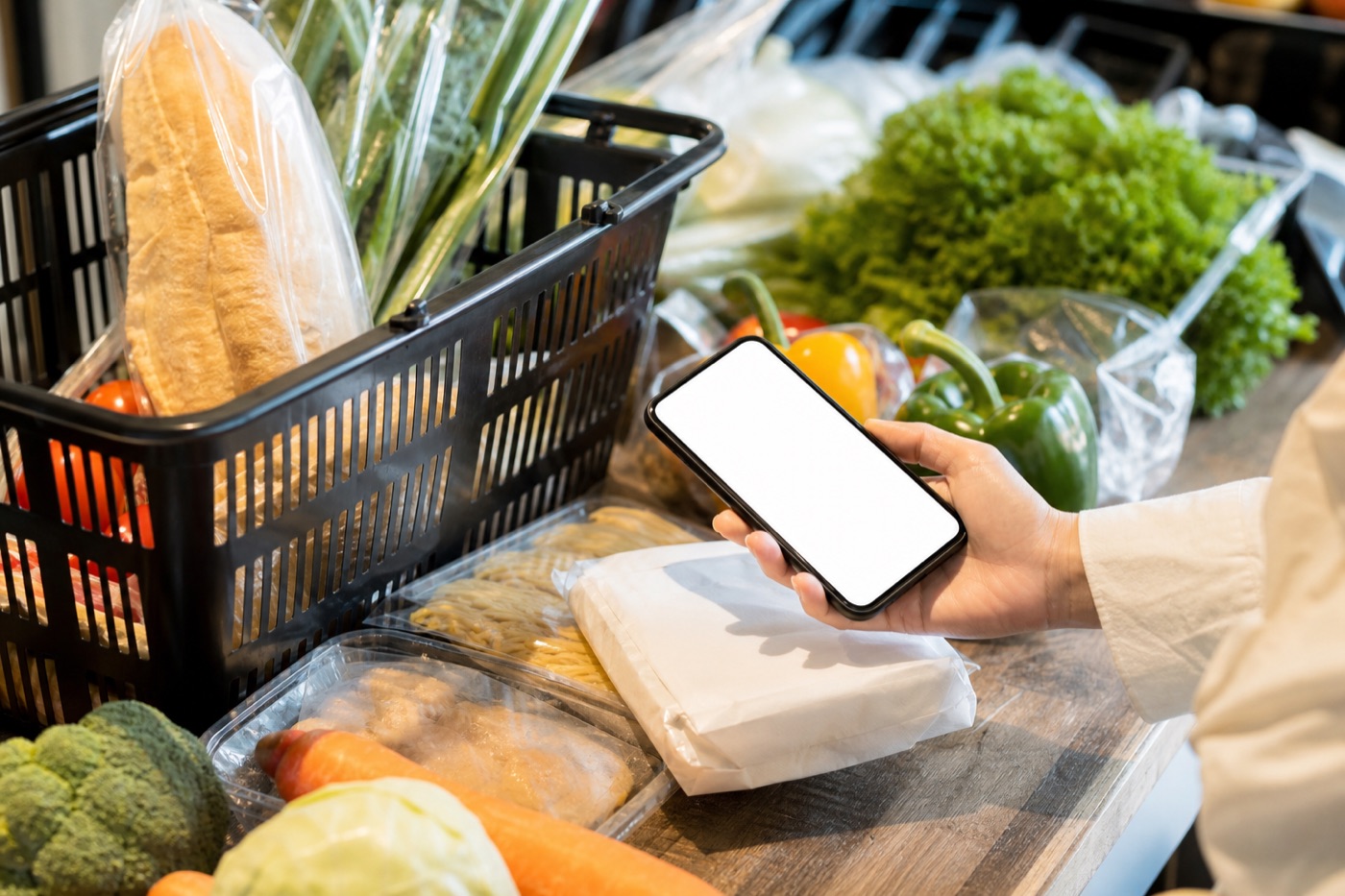 A grocery basket with packaged foods and fresh vegetables, and a hand holding a phone above a package